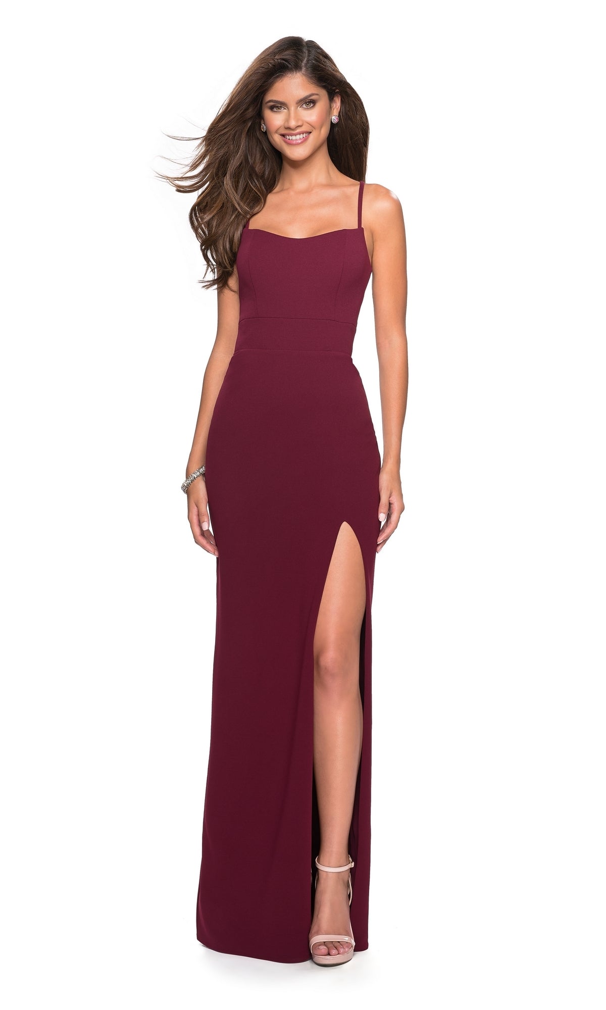 A woman stands smiling against a white background in the La Femme Strappy Open-Back Jersey Prom Dress 26940—a sleeveless, floor-length burgundy gown with an open back and thigh-high side slit—paired with strappy heels.